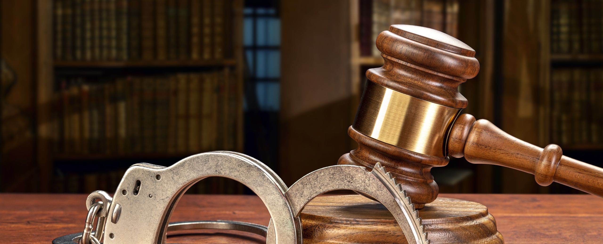 A judge’s gavel and a pair of handcuffs on a desk in front of a law library
